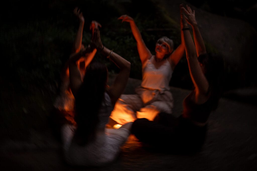 Frauen sitzen auf einem Stein mit Kerzen in der Mitte, heben ihre Hände nach oben in Schwesternschaft, sind frei und wild